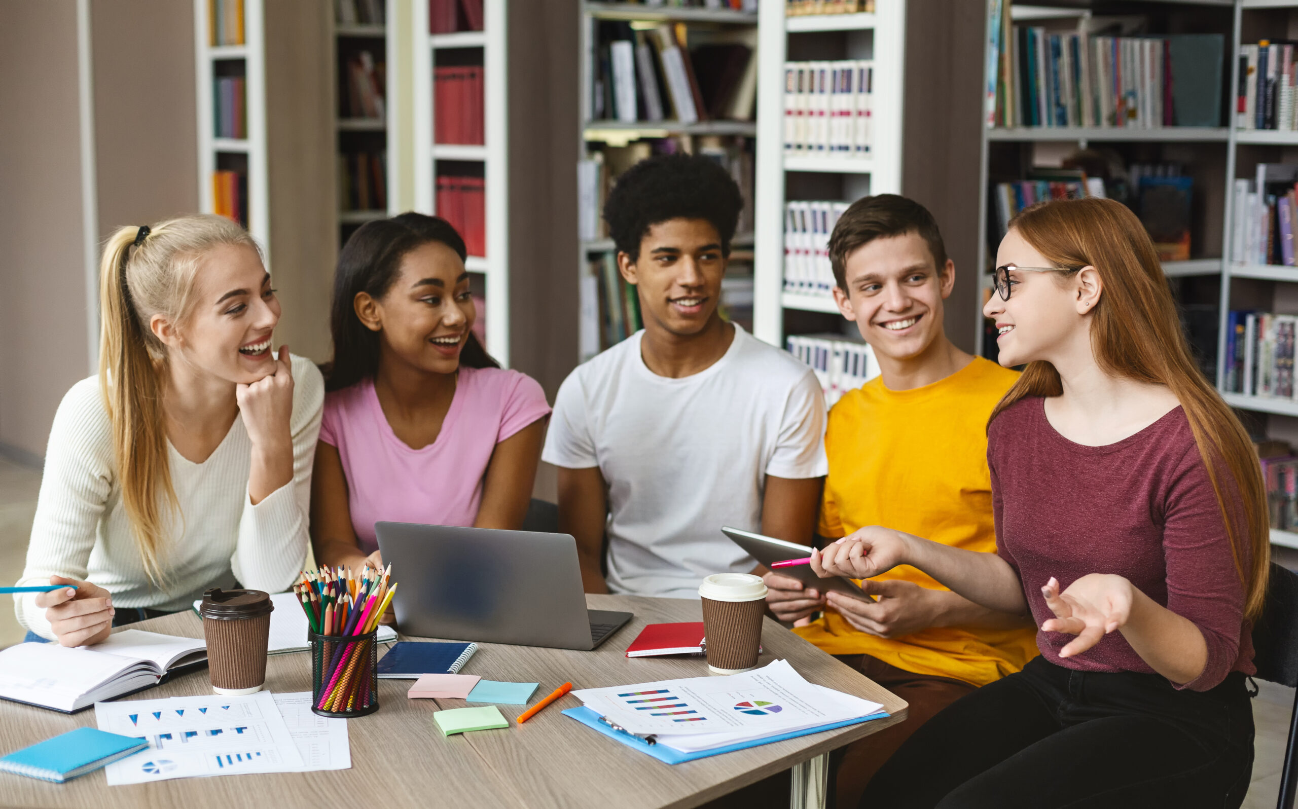 Group of diverse students collaborating around a table with laptops, charts, and notebooks in a library setting